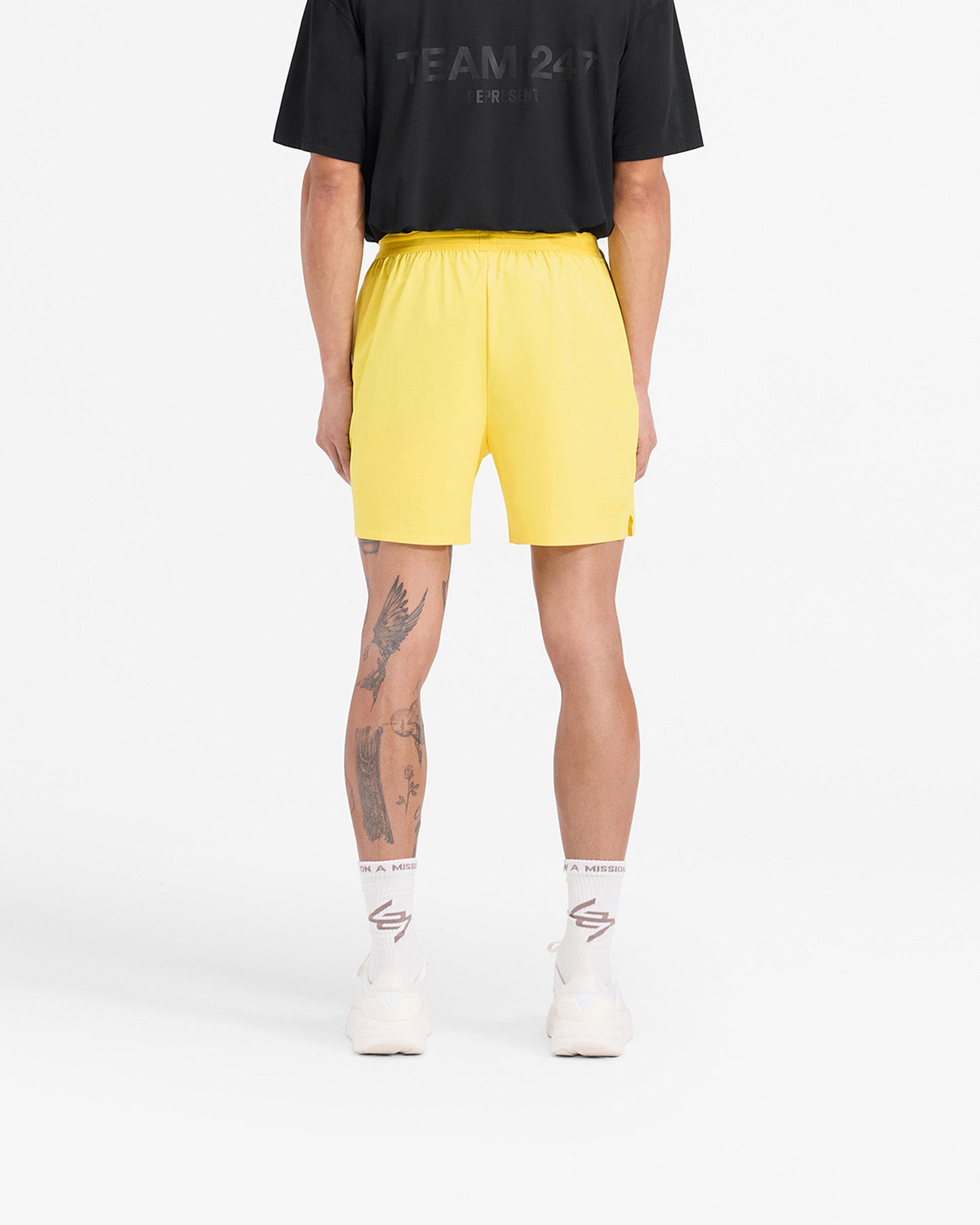 Man wearing yellow shorts on a plain white background | Team 247 Fused Shorts in Lemon | Gym shorts with quick drying fabric and 247 branding | 247 Shorts | Represent Clo